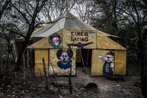 A dull yellow circus tent on which three clowns are painted along with Spiderman in a dismal wood